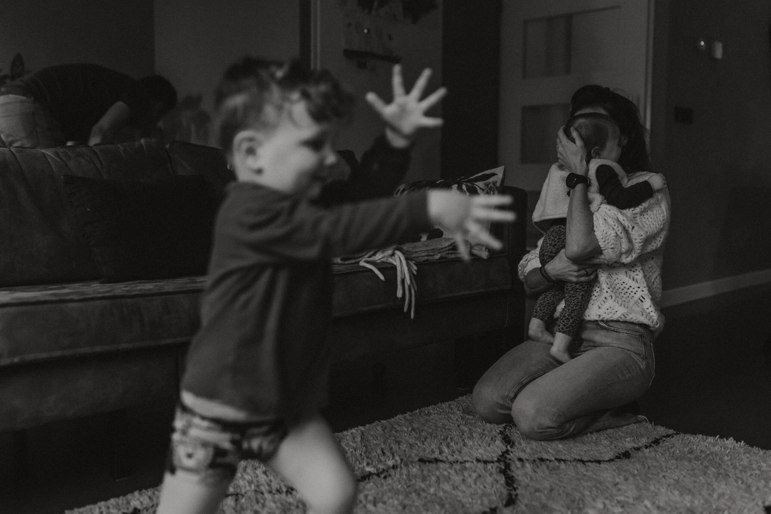 boy running through a living room with arm in front of him, while an adult women cuddles a kid in the background.
